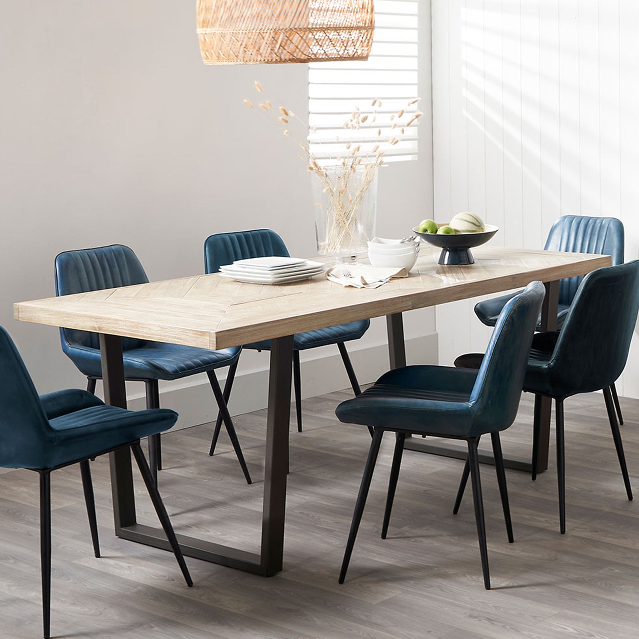 A modern dining room showcases the Pacific Lifestyle - Marca Acacia Wood Dining Table with industrial legs, blue upholstered chairs, a fruit bowl, and a wicker pendant light, all exuding sleek Scandi styling.