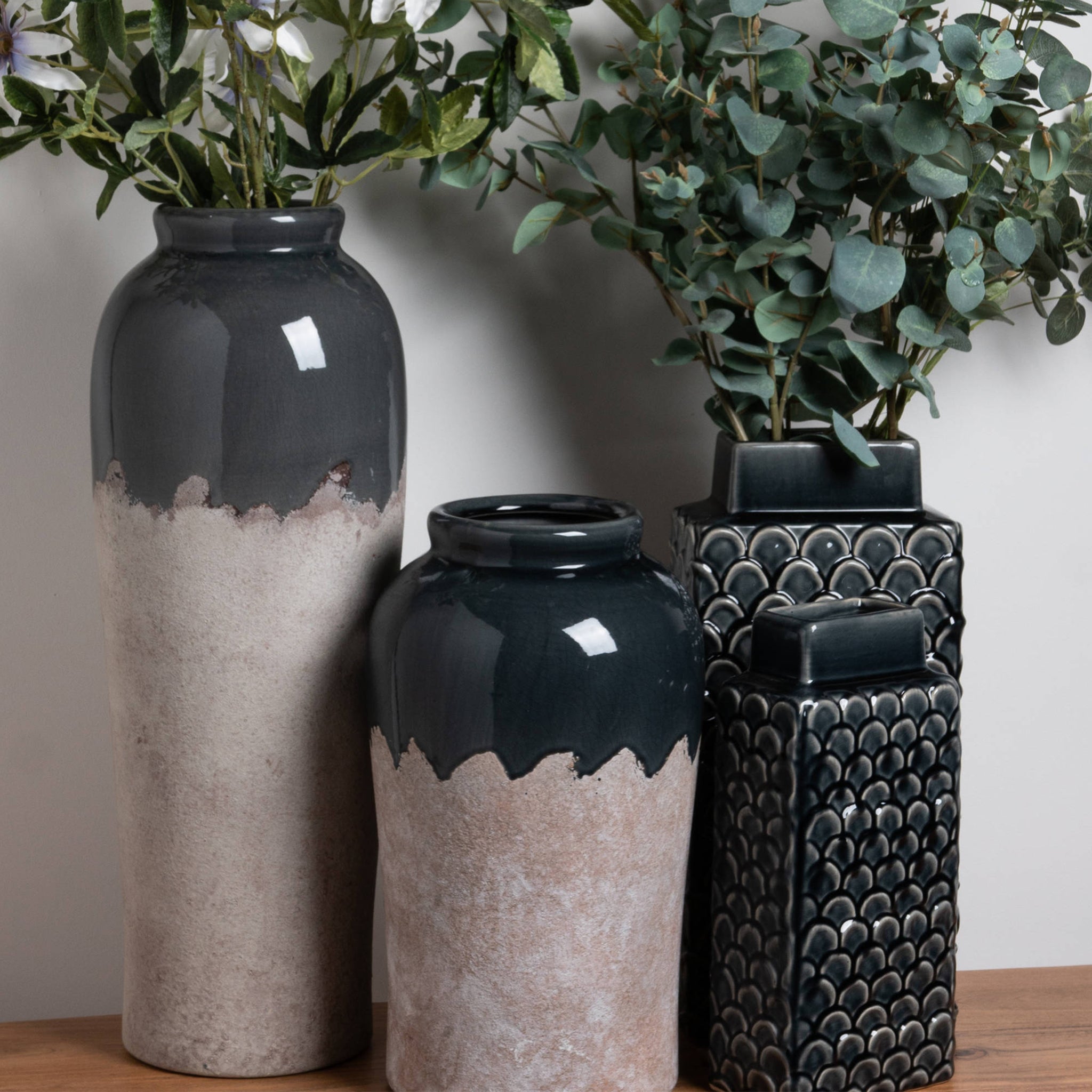 Three decorative vases with foliage on a wooden surface, showcasing black and beige tones and diverse textures, including the Medium Conran Vase, which is ideal for long stem floral arrangements.