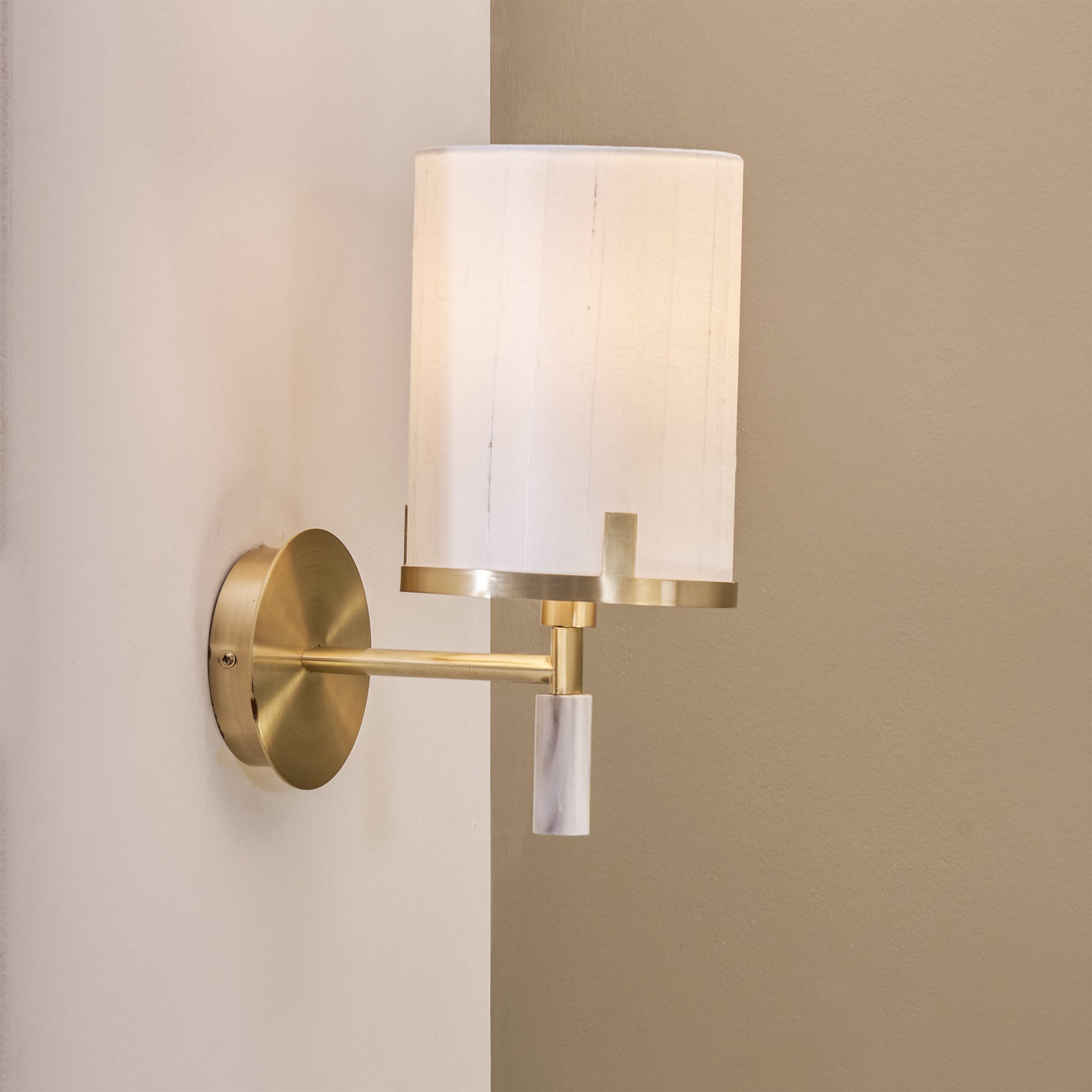 The Pacific Lifestyle Midland Champagne Gold Metal and Marble Effect Wall Light, featuring a white cylindrical lampshade, is mounted on a light tan wall, bringing hotel-style elegance with its modern design.