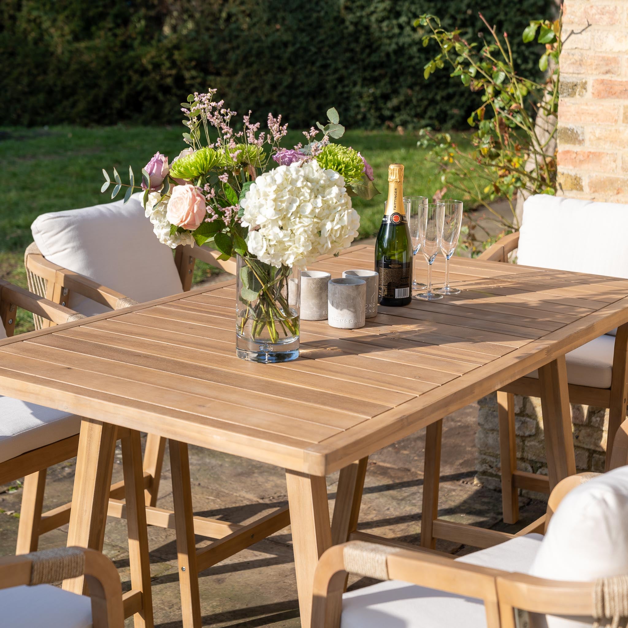 A Quay 6 Seat Rectangular Bar Set in Linen, featuring a wooden outdoor table made from sustainably sourced acacia wood, adorned with a bouquet of flowers, a bottle of champagne, two glasses, and surrounded by cushioned chairs.