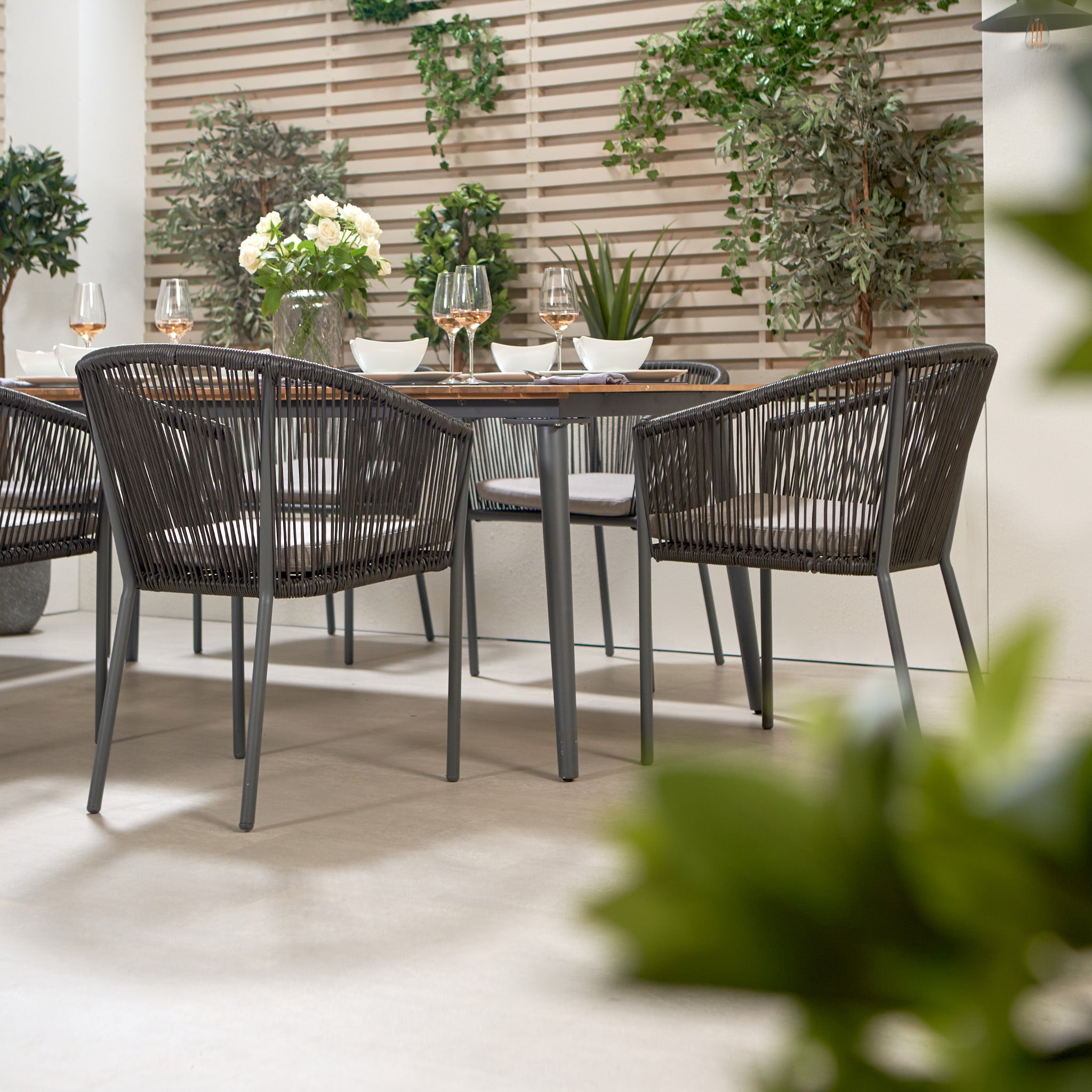 The outdoor patio boasts a Pacific Lifestyle Reims Dining Set in Grey with an elegant Acacia wood-topped table, contemporary dining chairs, lush plants, and wooden paneling as the backdrop.