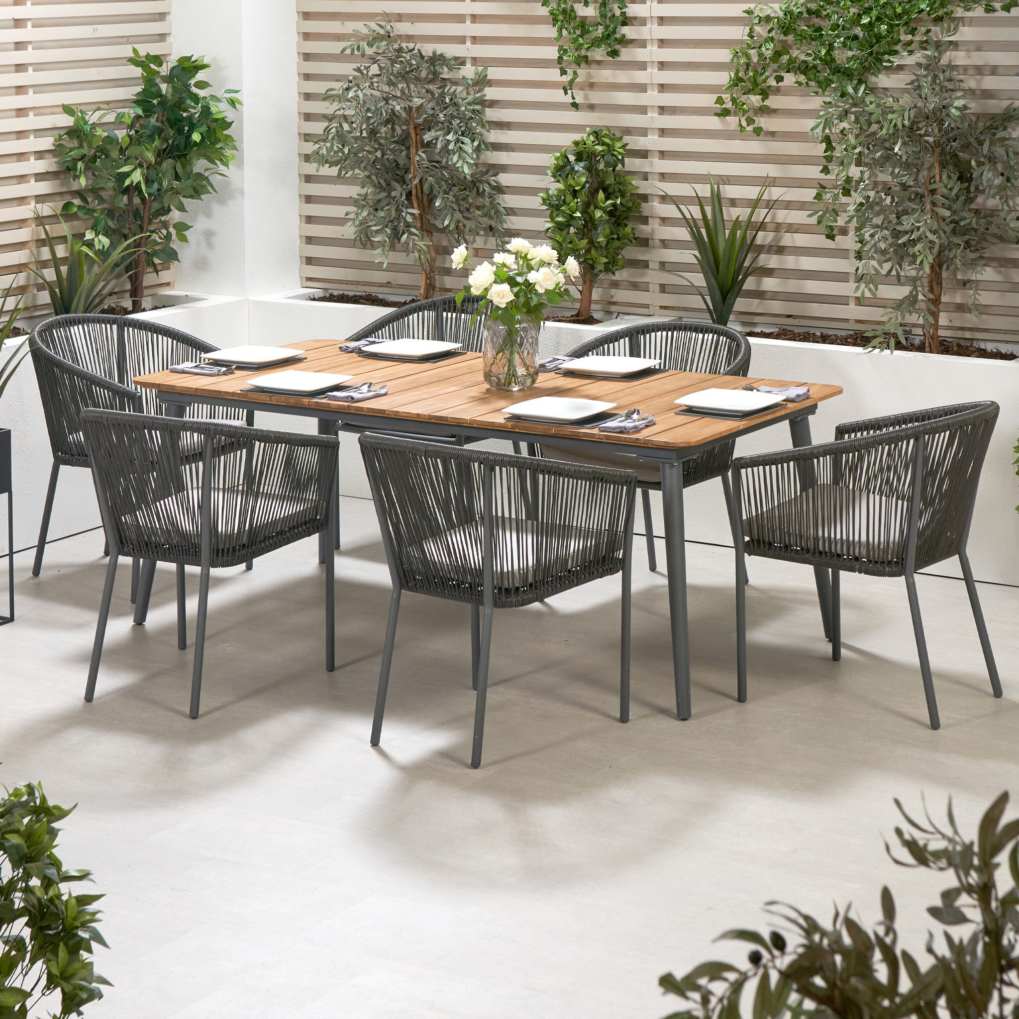 The Pacific Lifestyle Reims Dining Set in Grey includes acacia wood-topped table and contemporary chairs, beautifully accented by potted plants and white lattice panels.