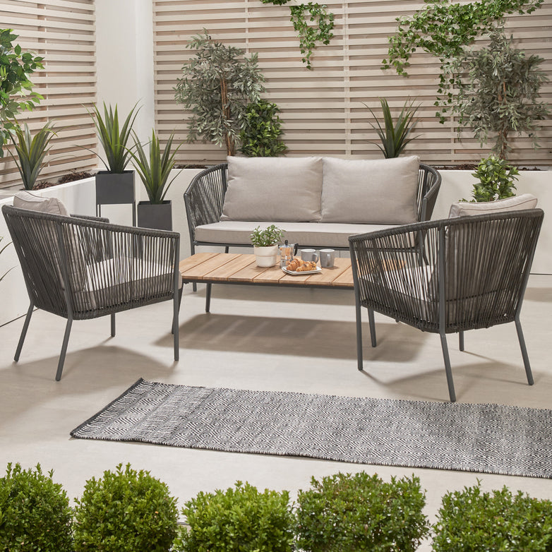 The Pacific Lifestyle - Reims Lounge Set Grey includes a chic modern outdoor patio arrangement with an acacia wood sofa, two chairs, a wooden table, lush plants, and polystring slatted privacy screens.