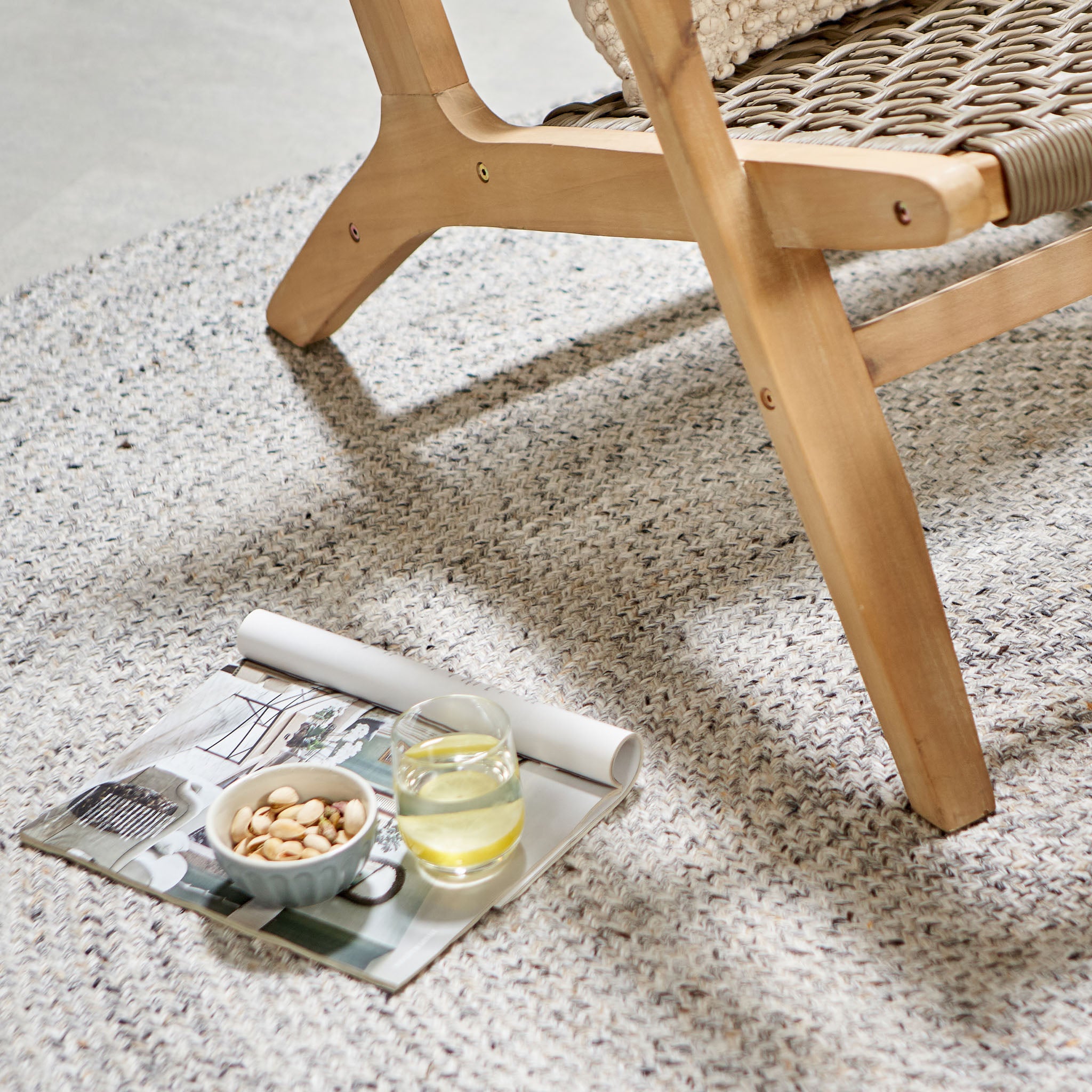 The Sesto Lounge Chair & Hocker Set by Pacific Lifestyle features woven chair legs on a rug, with a magazine, bowl of nuts, and a glass of yellow drink nearby. This Acacia wood garden furniture is ideal for leisurely afternoons, combining elegance with the comfort of waterproof seating.
