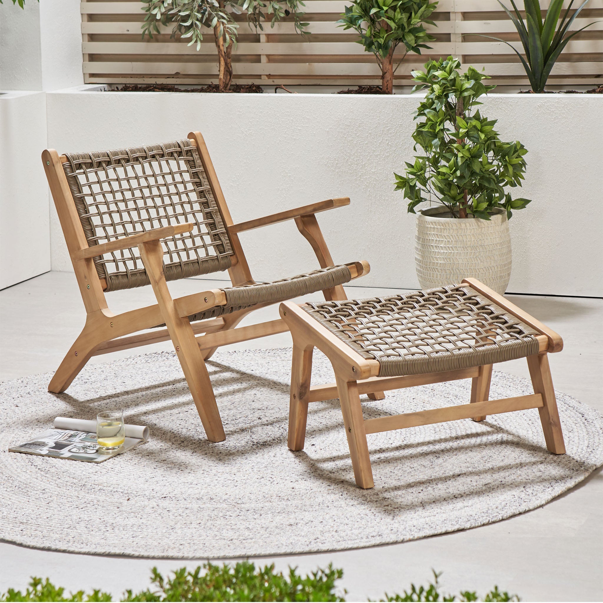 The Pacific Lifestyle Sesto Lounge Chair & Hocker Set, featuring acacia wood and woven waterproof seats, sits on a round rug in a modern patio setting surrounded by plants.