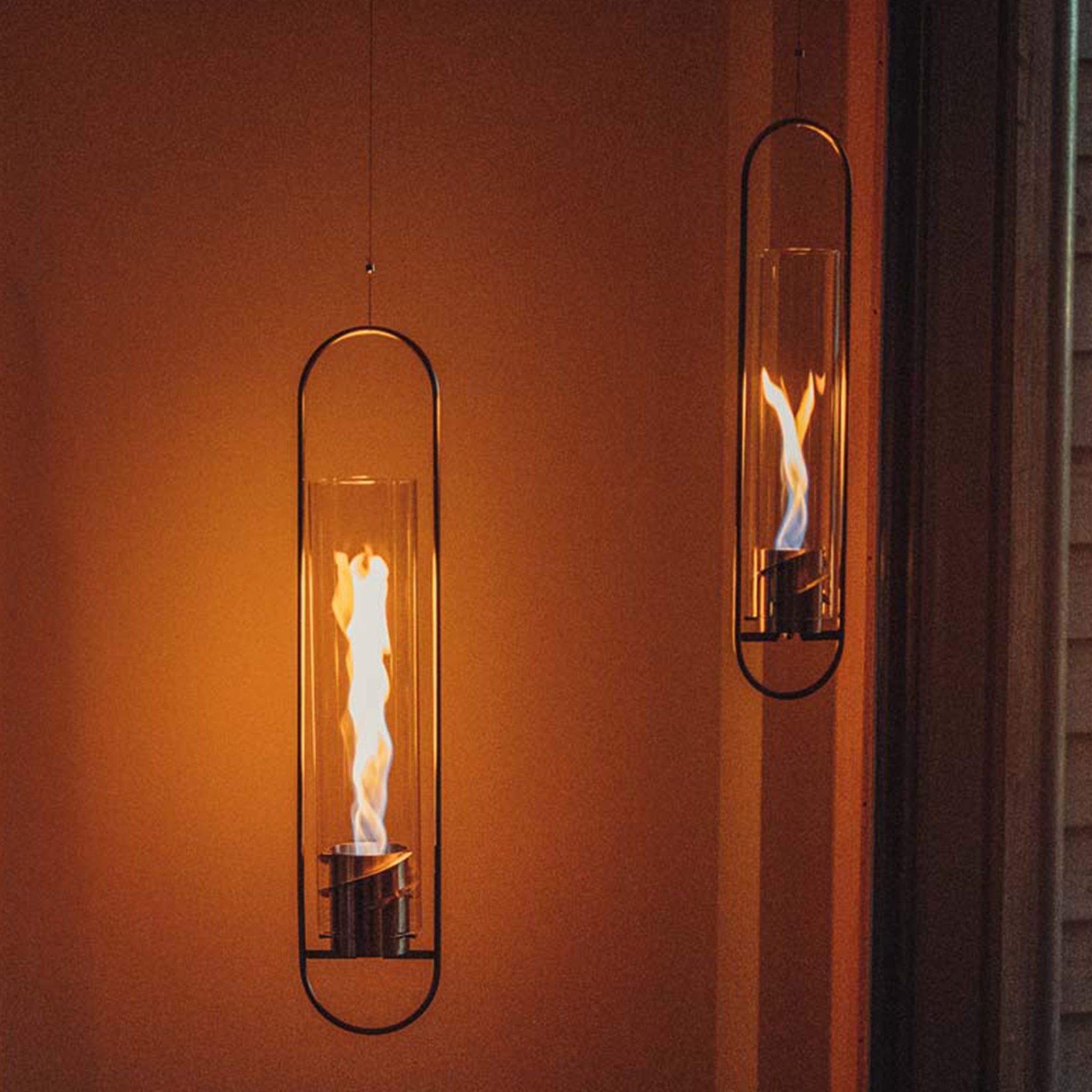 Two sleek Höfats SPIN 120 Hanging Fires in Silver with visible flames, casting a warm glow against a dimly lit wall.
