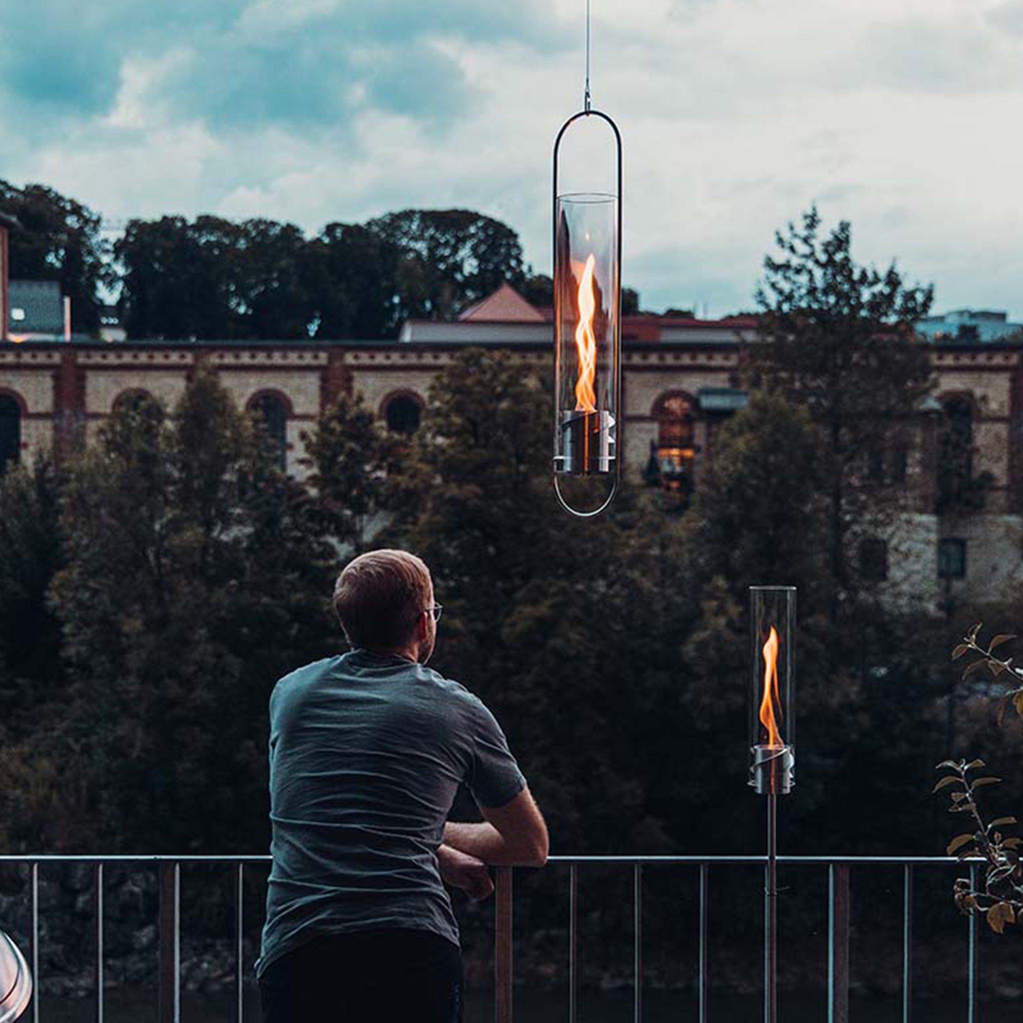 A person leans on a railing and gazes at two Höfats SPIN 120 Hanging Fires in Silver, with flames flickering, set against a backdrop of trees and buildings.