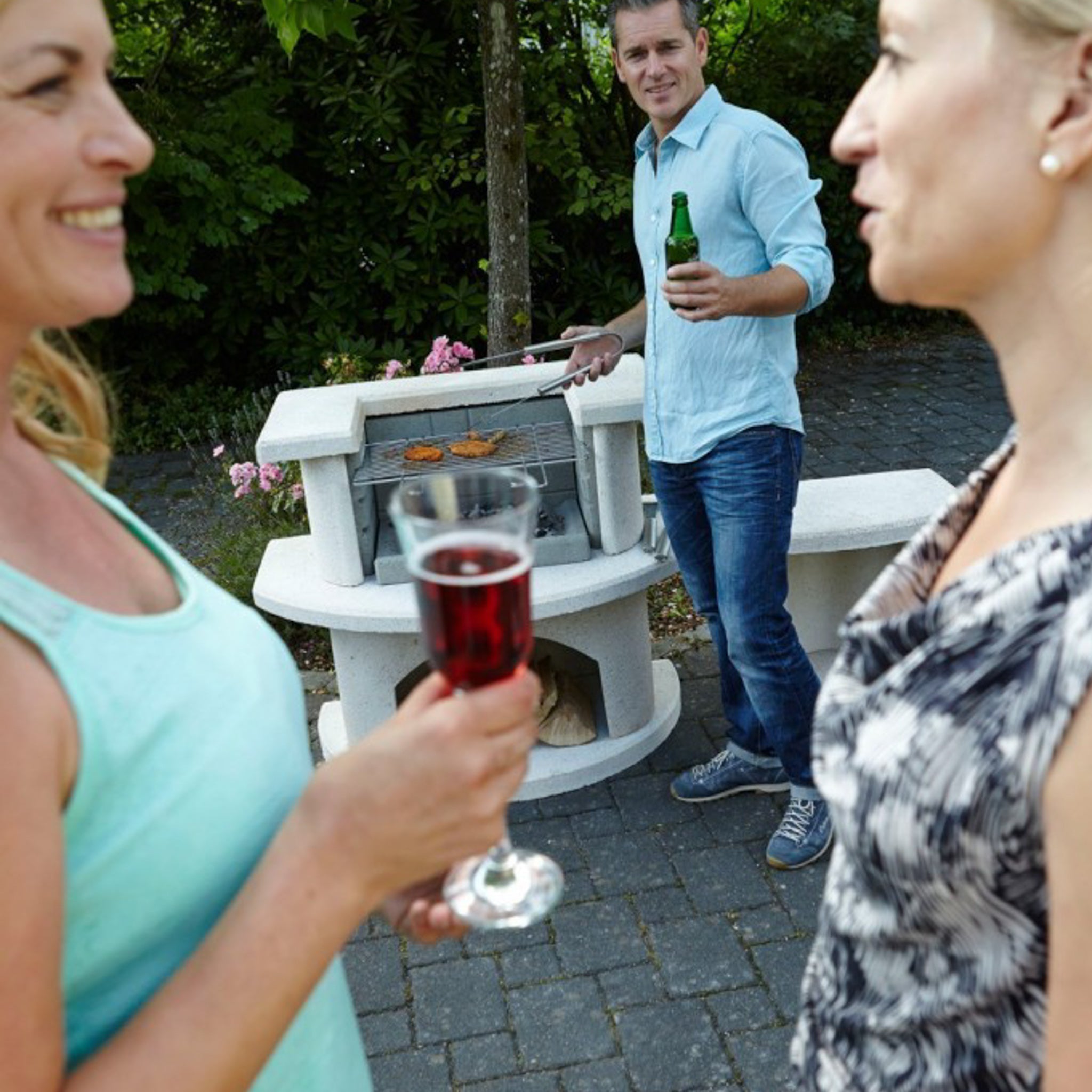 Two women chatting with drinks while a man grills food on a Buschbeck Venedig Grill Bar Masonry Barbecue in a garden setting.