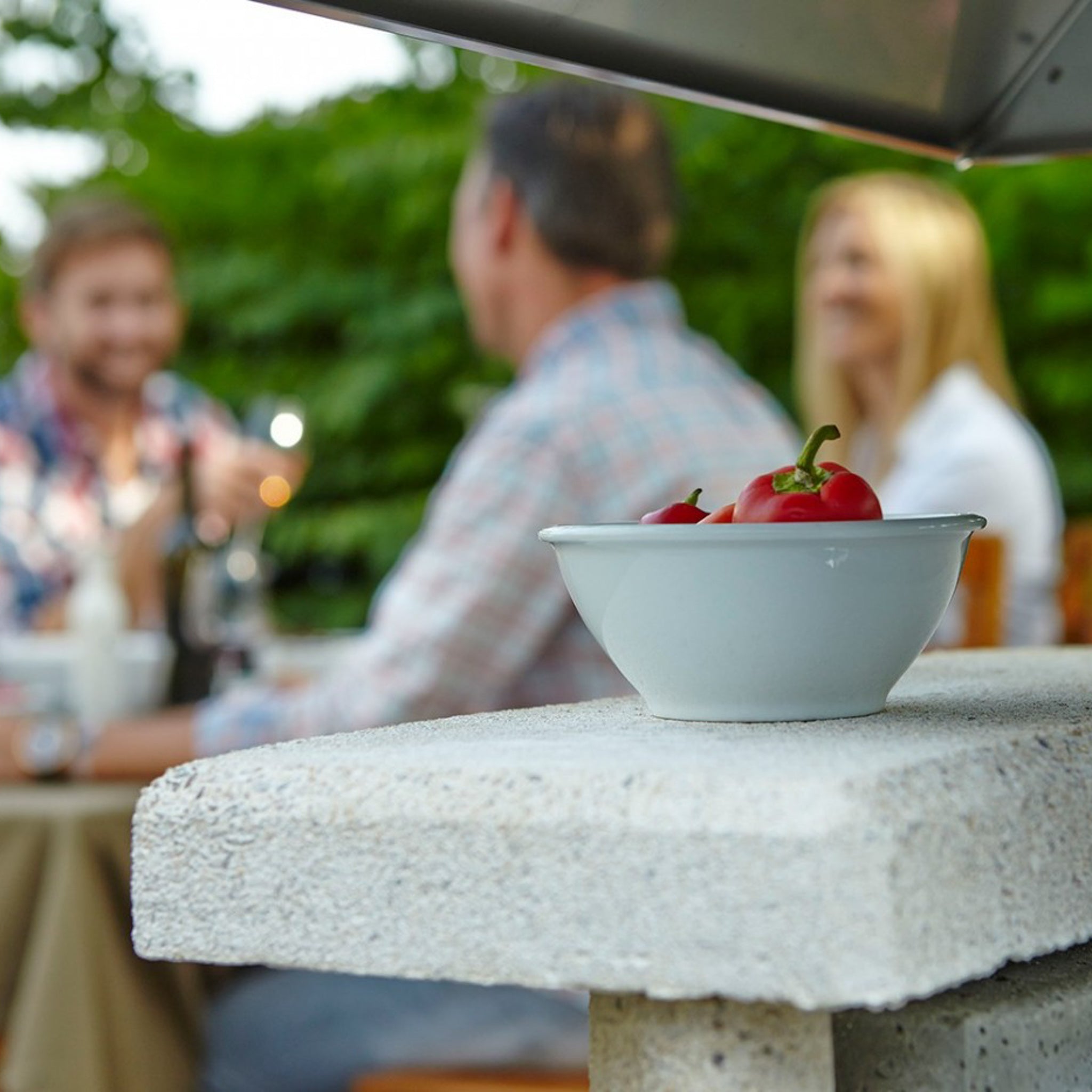 A bowl of red peppers rests on a stone ledge next to the Buschbeck Zurich Masonry Barbecue, while three people dine at an outdoor table in the background.